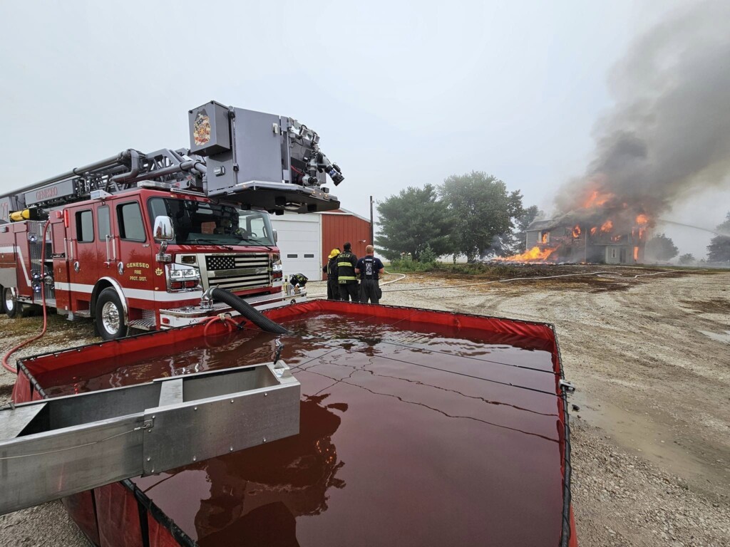 Firefighters using a FOL_DA_TANK portable folding take to put out a housefire that is happening in the background
